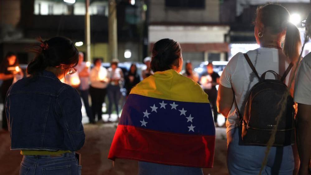 Relatives of political prisoners hold candles and posters during a vigil in Zamora, Venezuela, 26 February 2026. A woman, with her back to the camera, has a Venezuelan flag wrapped around her shoulders. Others hold candles.