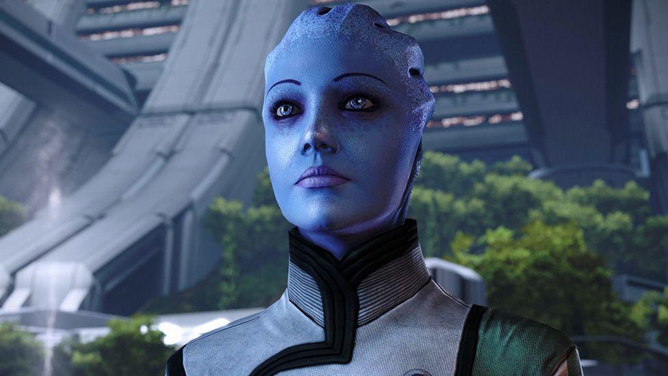 A screenshot from Mass Effect Legendary edition shows a blue, android character inside a spacious, futuristic atrium filled with trees. The grey, metallic structure that makes up the building is also visible in the background.