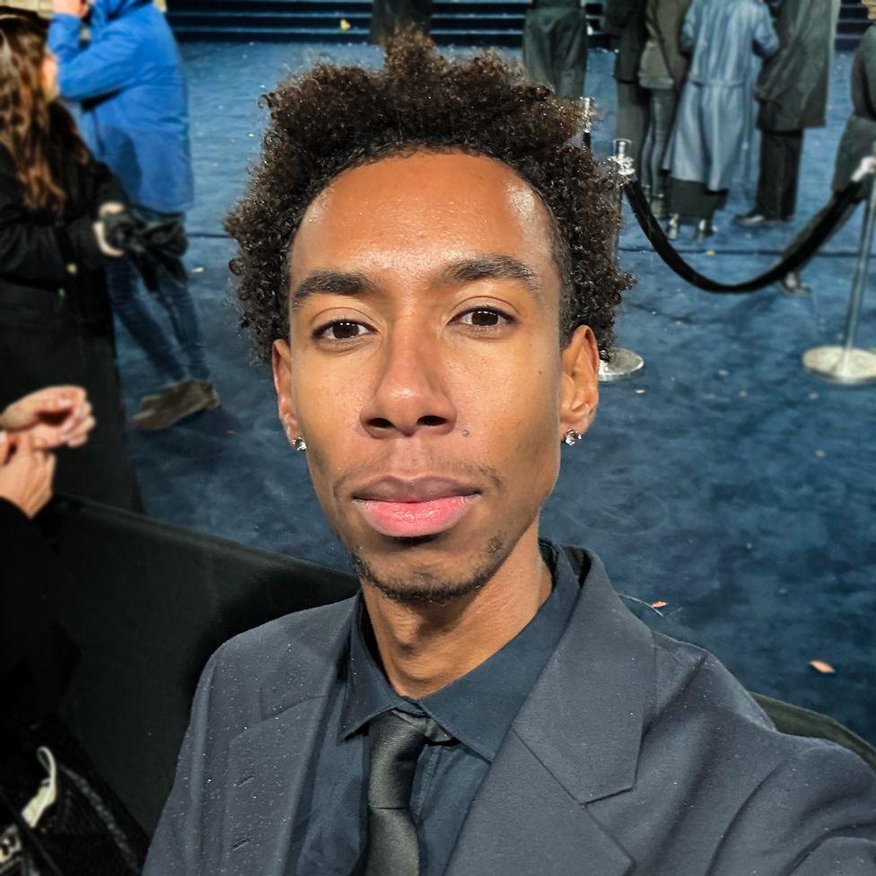 Joel is smartly dressed in a navy suit with a black tie and poses for a selfie in front of a blue carpet. Behind him are velvet ropes for a VIP area with people waiting in the background.