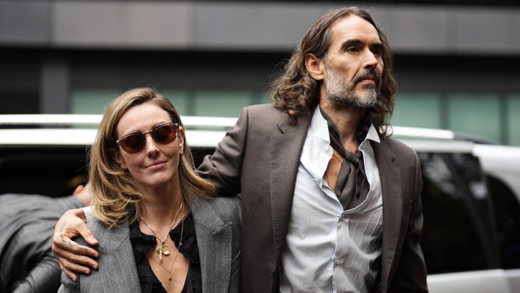 Russell Brand walking in to court with one arm around his wife's shoulder, she has a grey suit jacket on while he is earing a brown suit jacket and matching cravat
