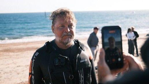 A man in an orange and black wetsuit poses for a photo on a beach with the sea behind him.