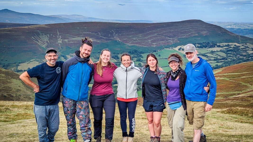 Seven people in outdoor clothing standing on top of a hill in the Brecon Beacons. Large and dramatic hills span the background.