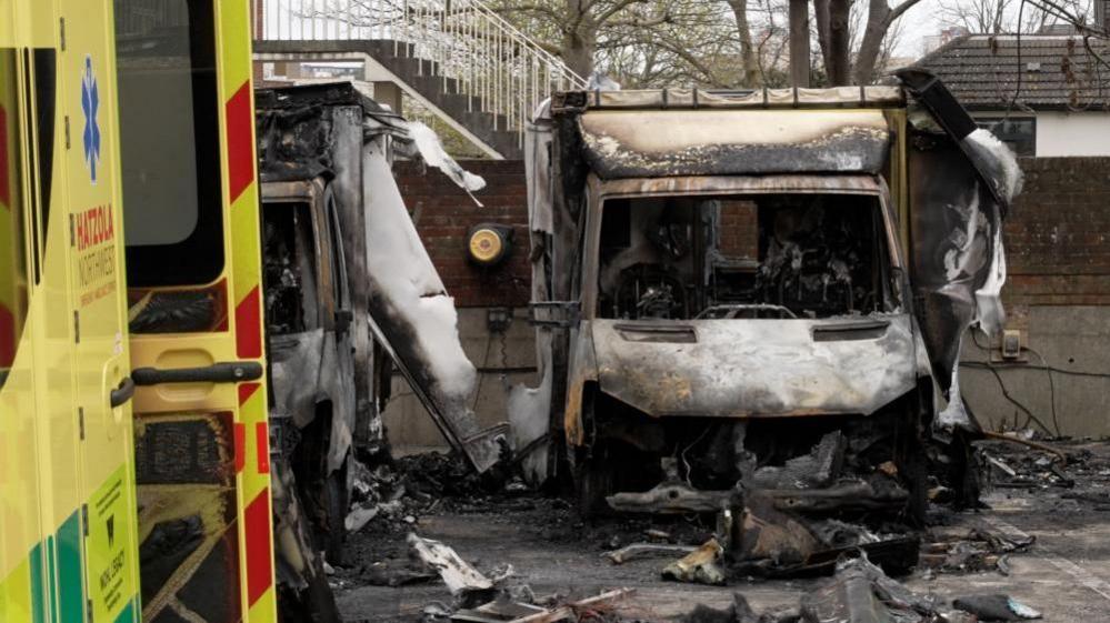 The burnt wreckage of an ambulance is seen in the right hand side of the image, where another vehicle, which is less burned is situated on the right of the picture.