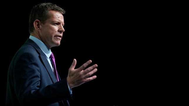 Rhun ap Iorwerth in a dark blue suit, light blue shirt and purple tie, his right arm is outstretched and he is speaking.
