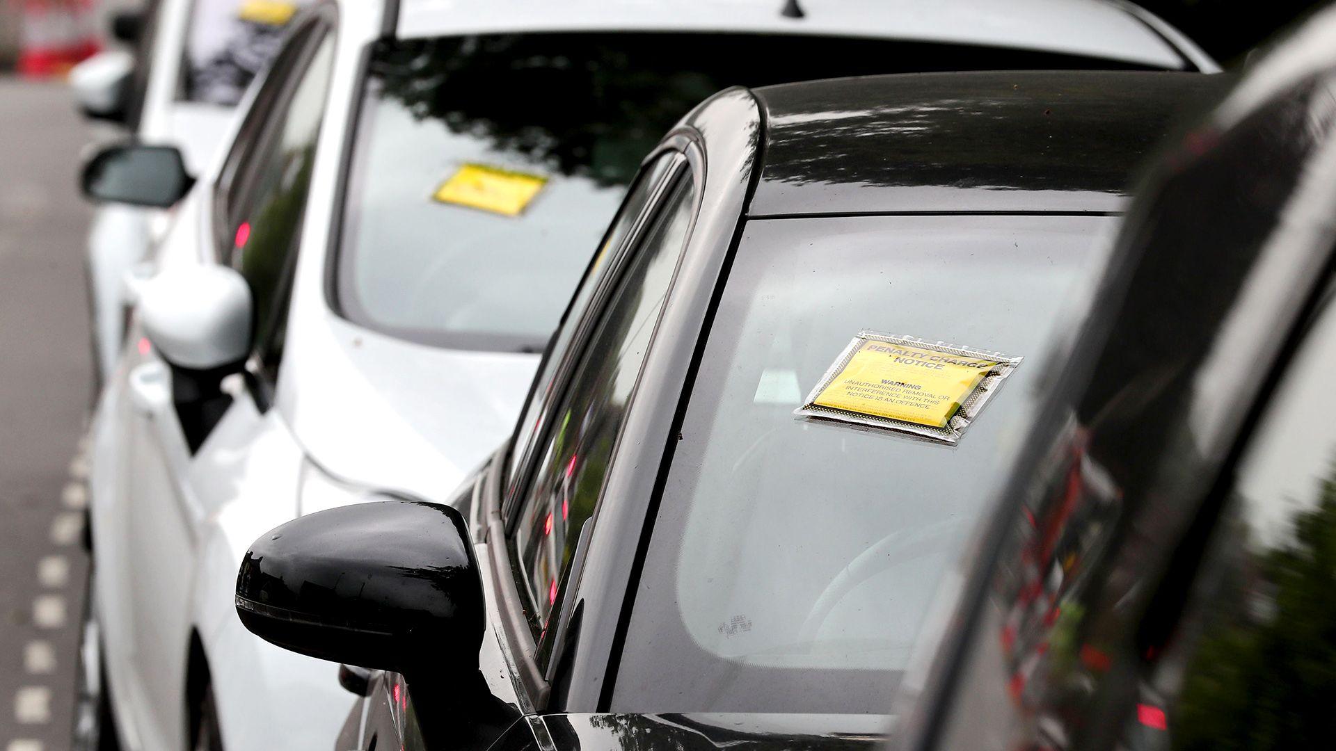 A generic-looking image shows a row of cars seen front-on with yellow and black Penalty Charge Notices affixed to their windscreens, on a road near Gatwick Airport in 2017.
