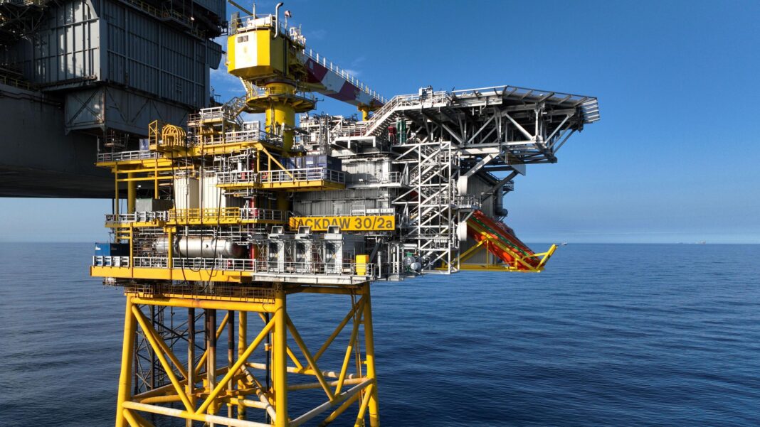 A mostly yellow-painted gas platform and allied grey rig stand in calm blue seas with a clear blue sky behind them. It is sunny.