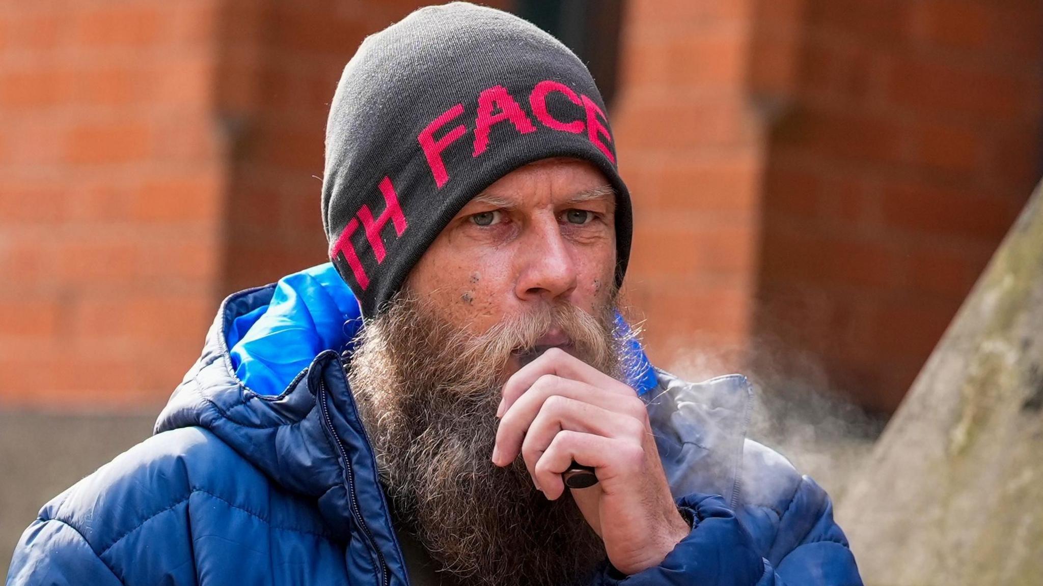 A man in his 30s, with a large beard, grey wool hat and blue bubble coat, stands outside a stone building holding a black vape to his mouth.