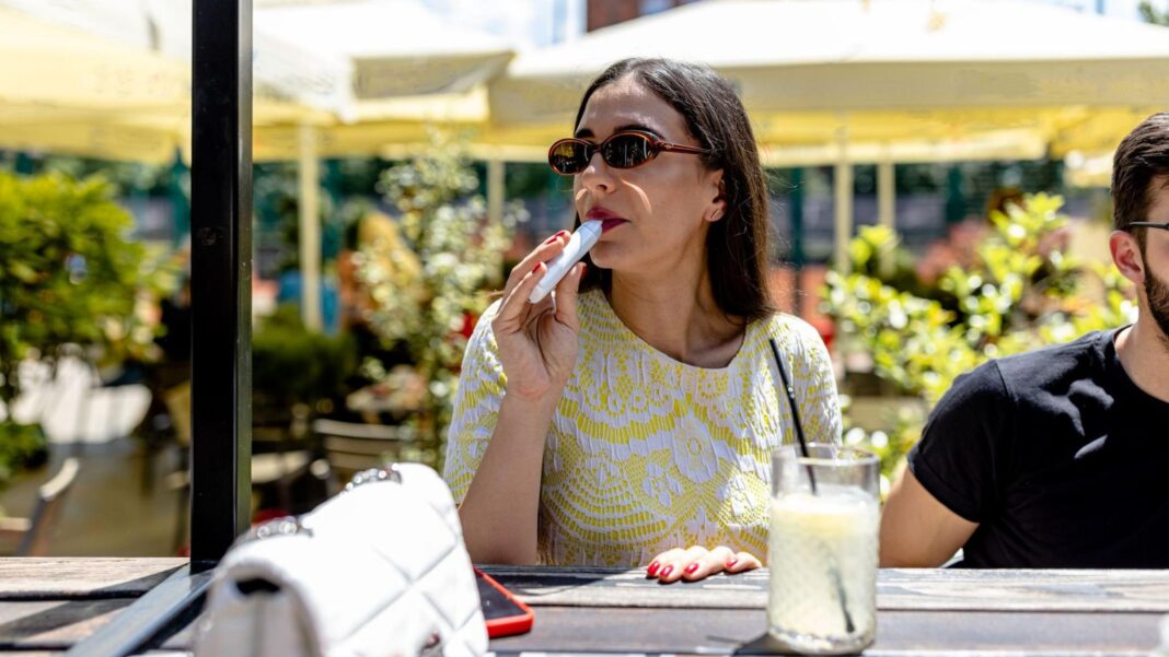 A woman vapes at an outdoor bar. She wears sunglasses and sits in the sun.