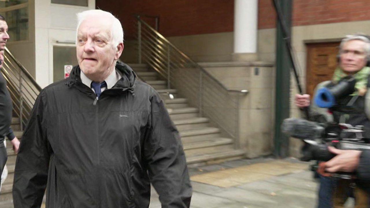 A man in his 70s with white hair, wearing a black coat, walks away from a court building with a camera crew filming him as he passes.