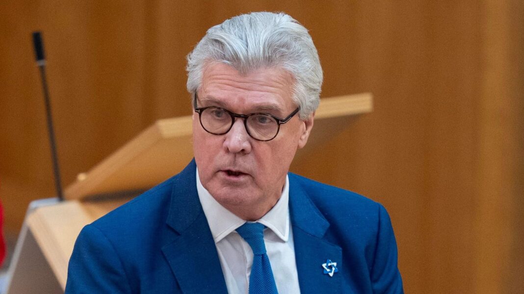 Malcolm Offord standing up in the Scottish Parliament. He is wearing a blue suit over a white shirt with a blue tie. He has white hair and is wearing glasses with a dark black frame.