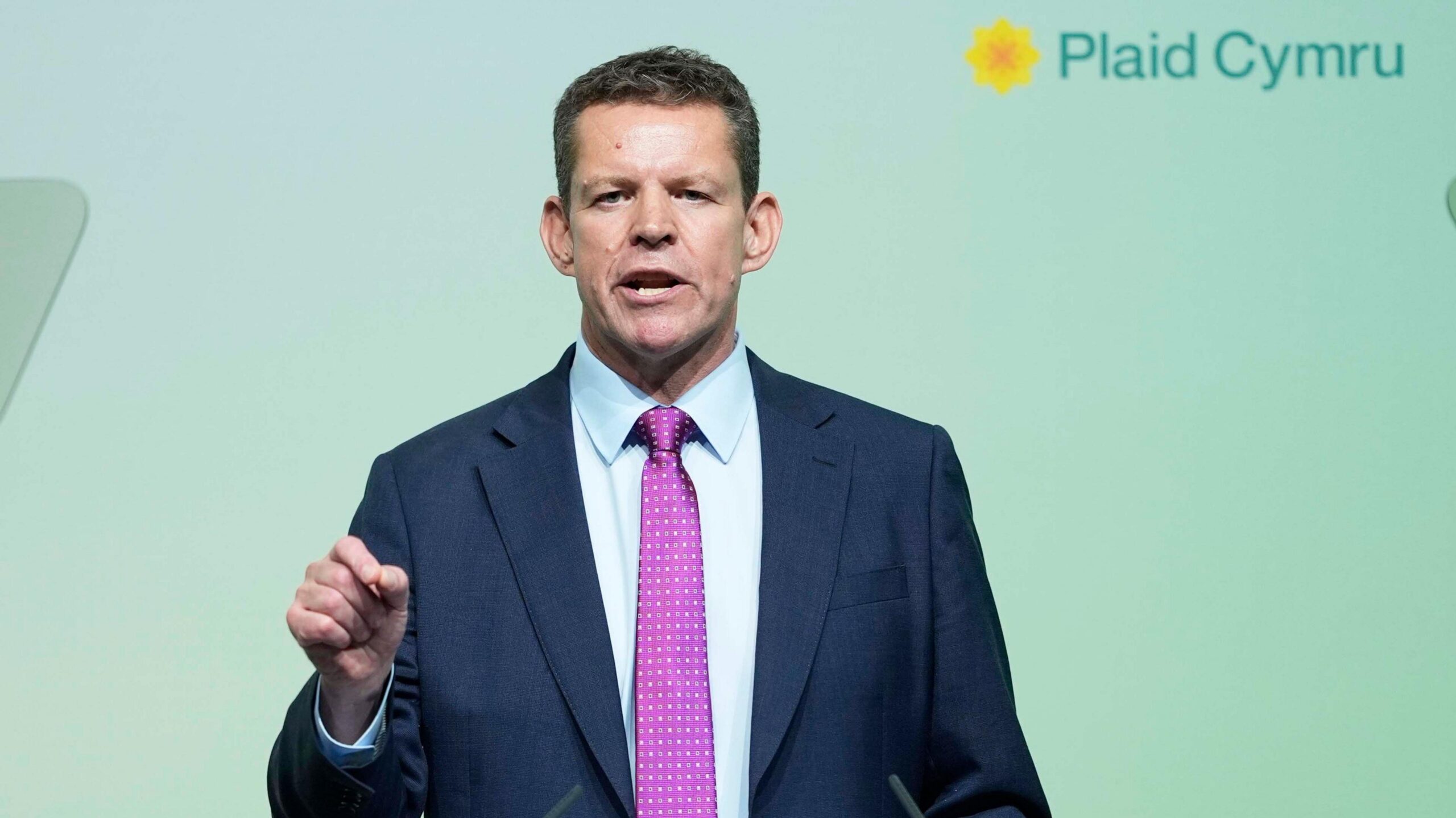 Rhun ap Iorwerth in a dark blue suit, light blue shirt and a purple tie. He is stood in front of a green background with the logo of Plaid Cymru.