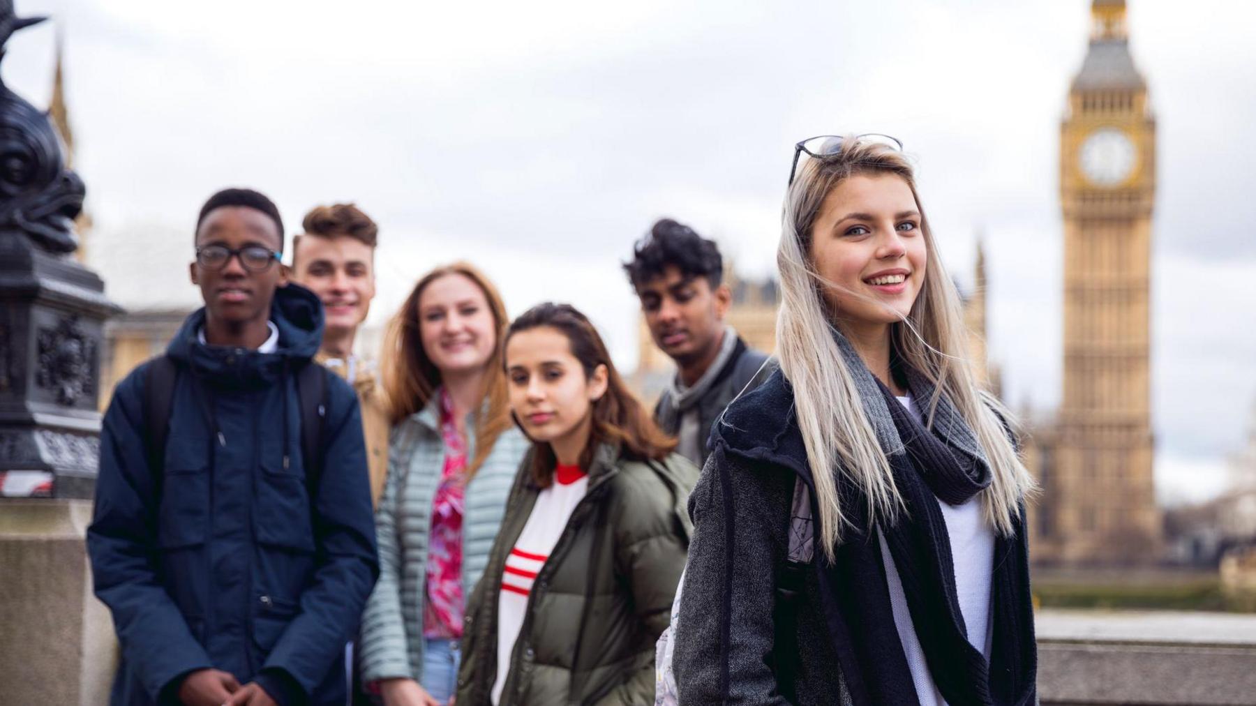 A group of six young people posing for a picture in front of the Palace of Westminster. At the forefront of the image is a girl with long blonde hair wearing a black jacket, white T-shirt, dark-grey scarf and glasses on the top of her head. Three boys and two girls, all dressed for autumn weather, stand behind her, slightly out of focus.