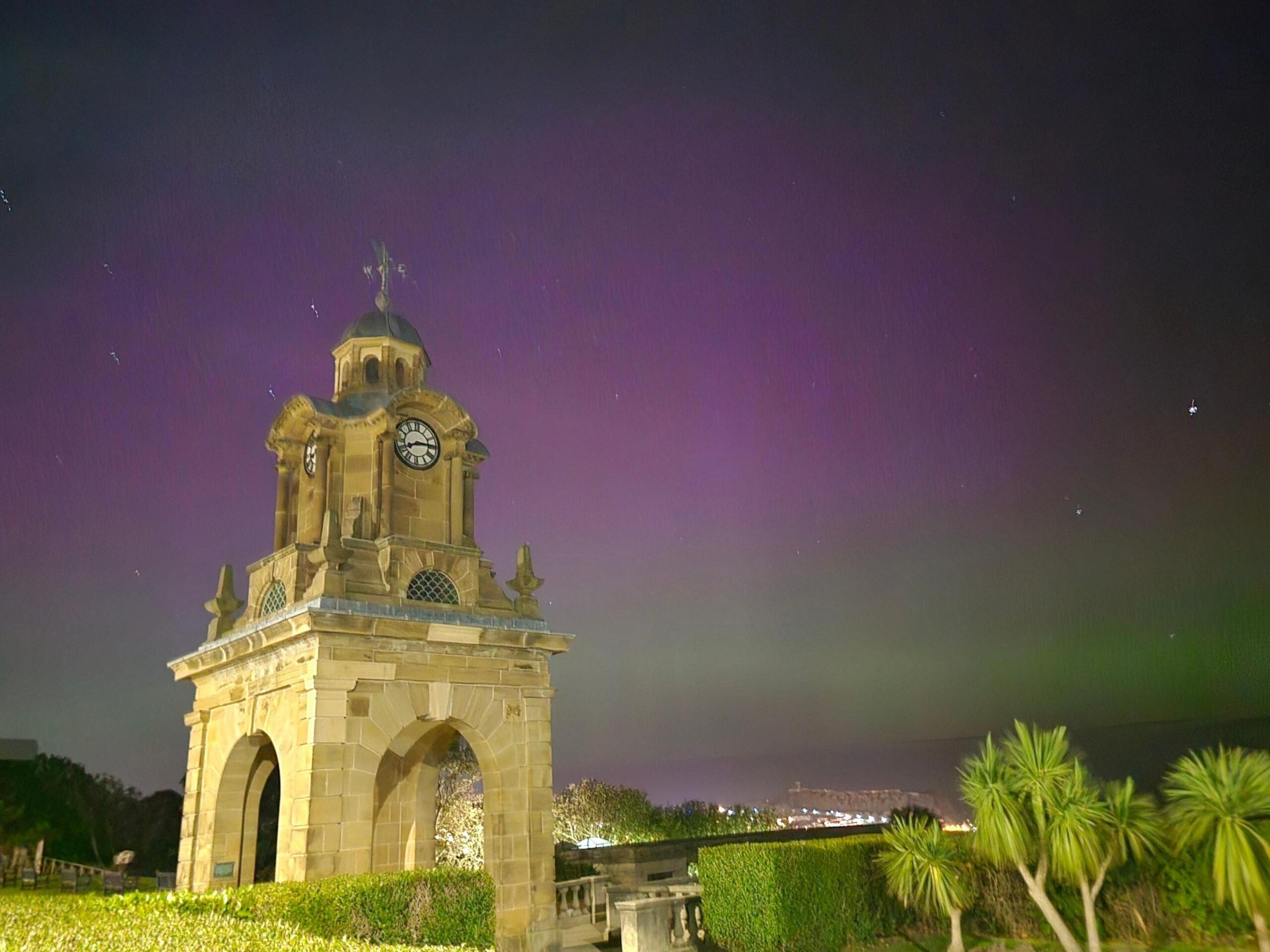 An image of the sky in Scarborough, North Yorkshire, on 10 October during the evening. The Northern Lights can be seen in a pink hue above a bell tower and green plants below.