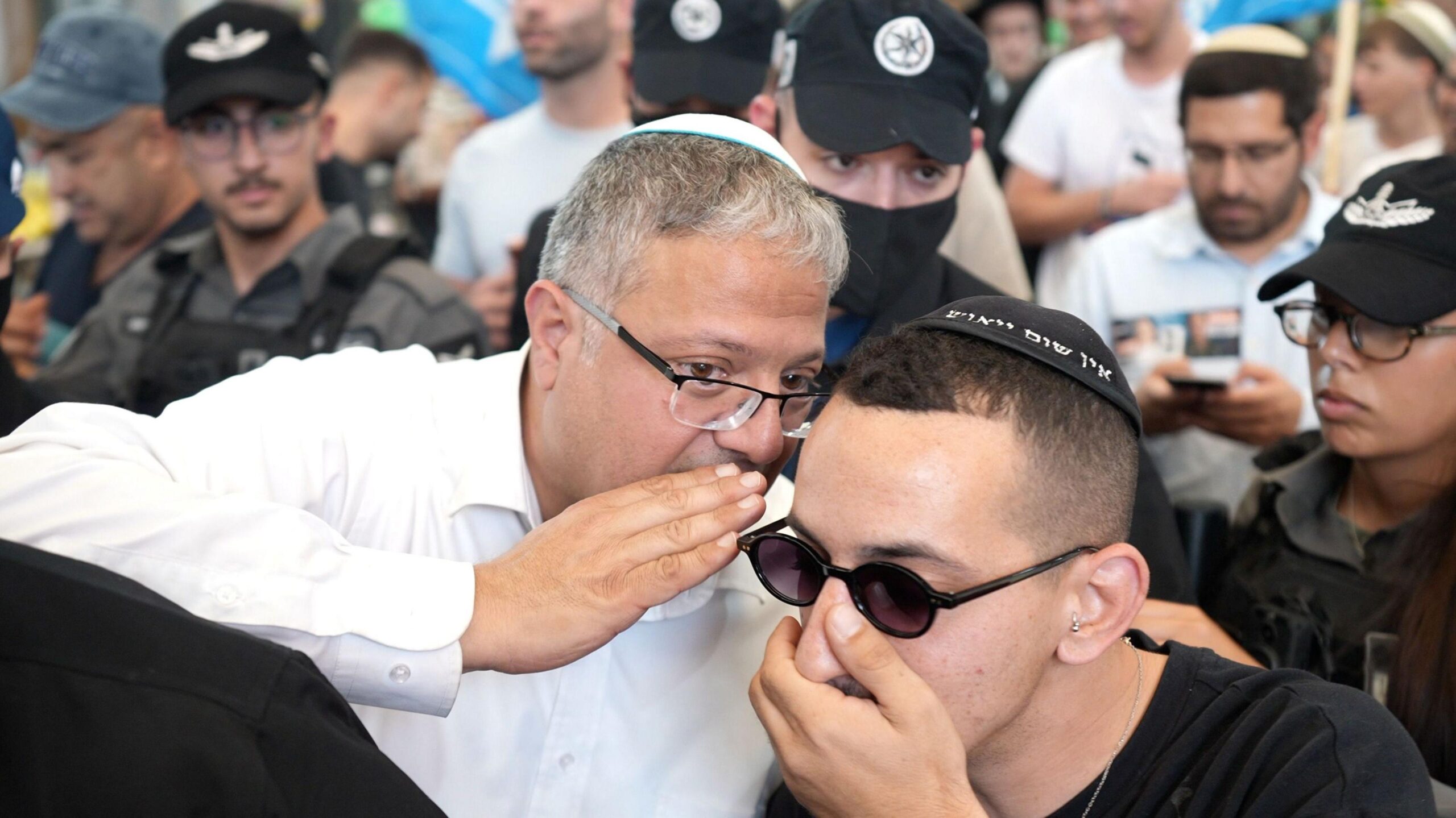 Itamar Ben-Gvir, wearing a white shirt and white skullcap, leans in to talk to another man, with several individuals behind them, some in protective gear