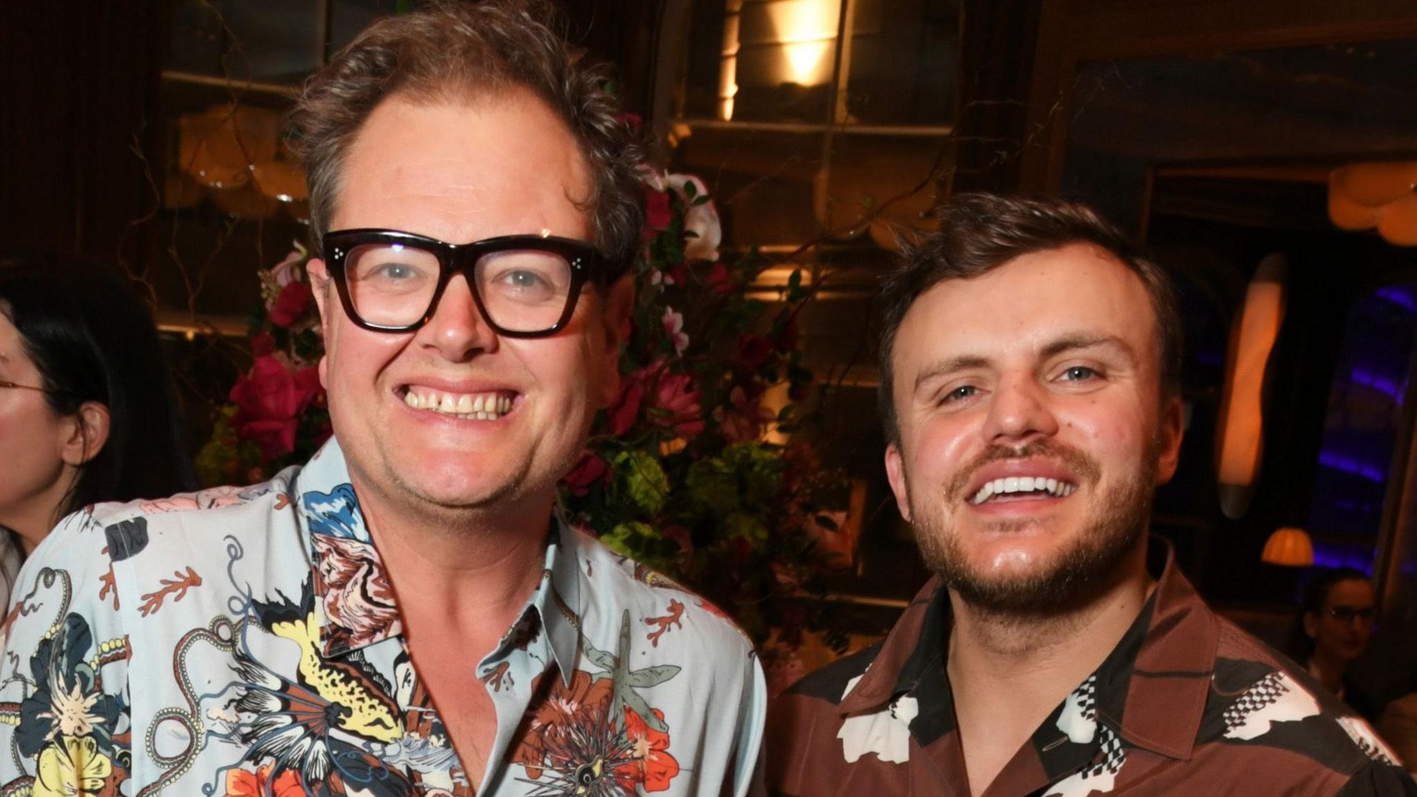 Alan Carr and Lee Peart standing together in patterned shirts and smiling at the camera in a party setting