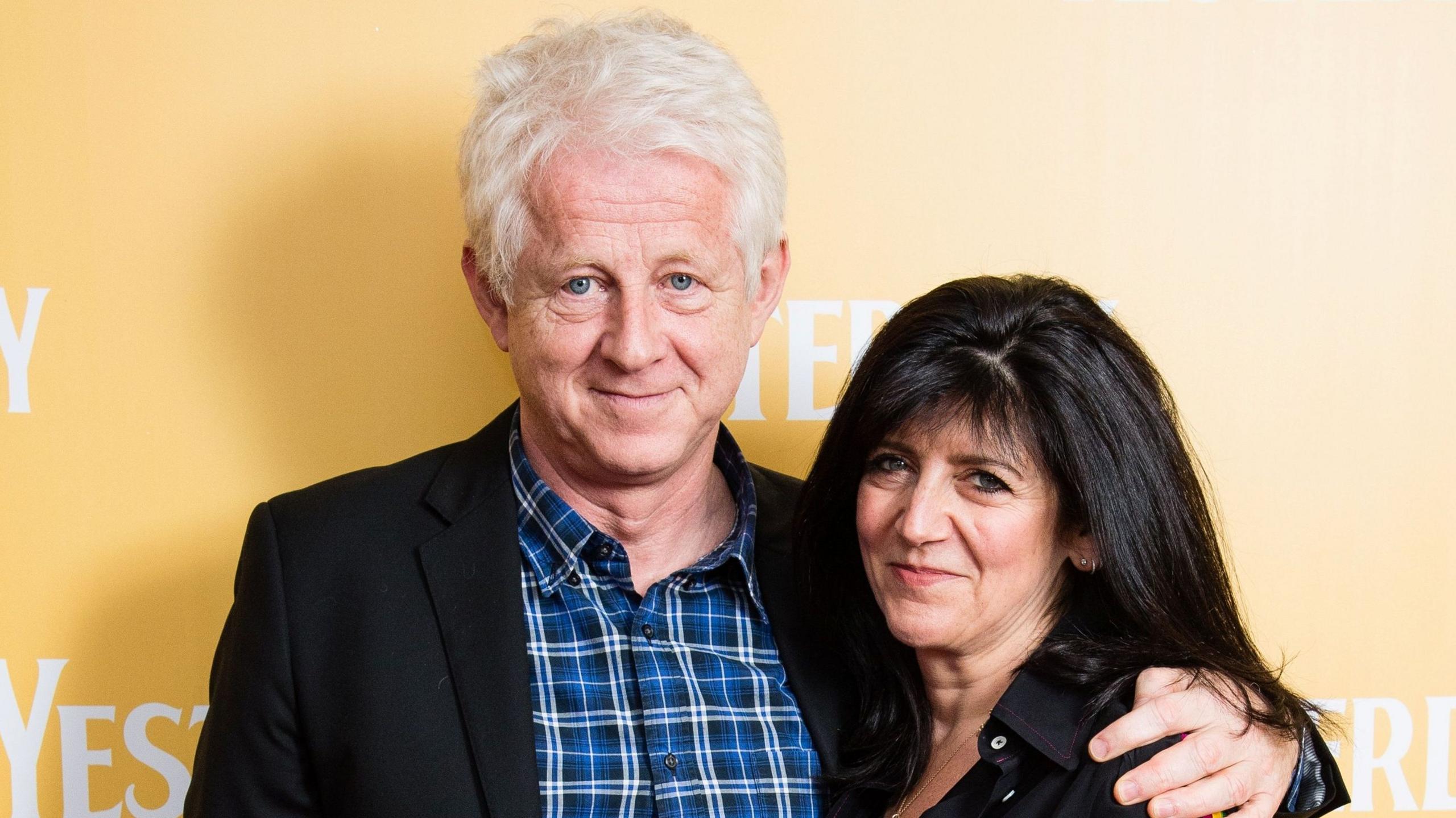 Richard Curtis and Emma Freud at a premiere for the movie, Yesterday. Curtis has his arm around Freud and they are both smiling for the camera, in front of a yellow backdrop with the logo for "Yesterday" written on it in white. Curtis has white hair and is wearing a chequered blue shirt and a black jacket. Freud has long black hair and is wearing a black jacket.