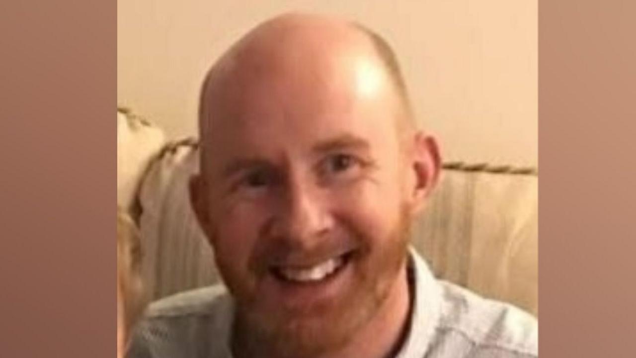A man smiling at the camera in a head and shoulders image. He has a bald head with very short ginger hair at the sides and a ginger beard. He is wearing a pale blue shirt and sitting on a cream sofa.