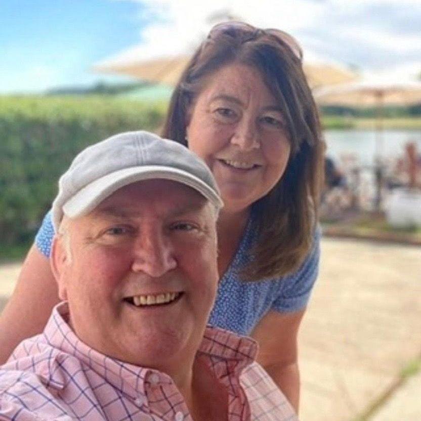 John is sitting down wearing a grey flat cap and Louisa is standing behind him with sand and beach umbrellas behind them