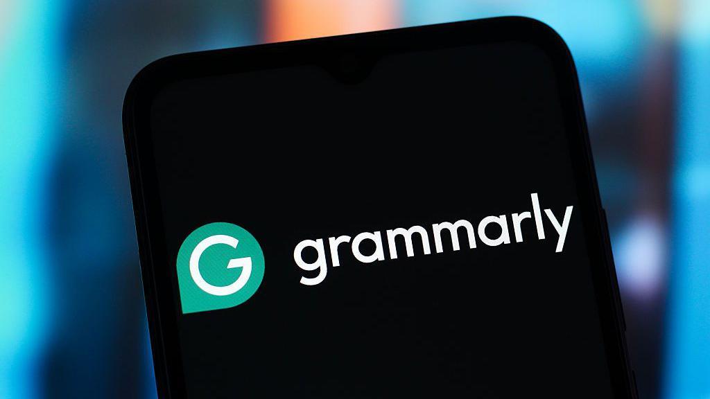 A close-up of a smartphone screen displaying the white and green Grammarly logo against a black background. The phone is held against a soft-focus, blue and orange bokeh background.
