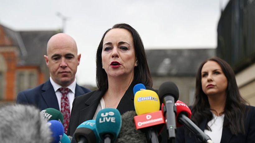A woman with long dark hair stands in front of a number of microphones. Behind her are two people. On the left is a bald man wearing a suit. On the right is a woman with long dark hair wearing a white top and a dark blazer.