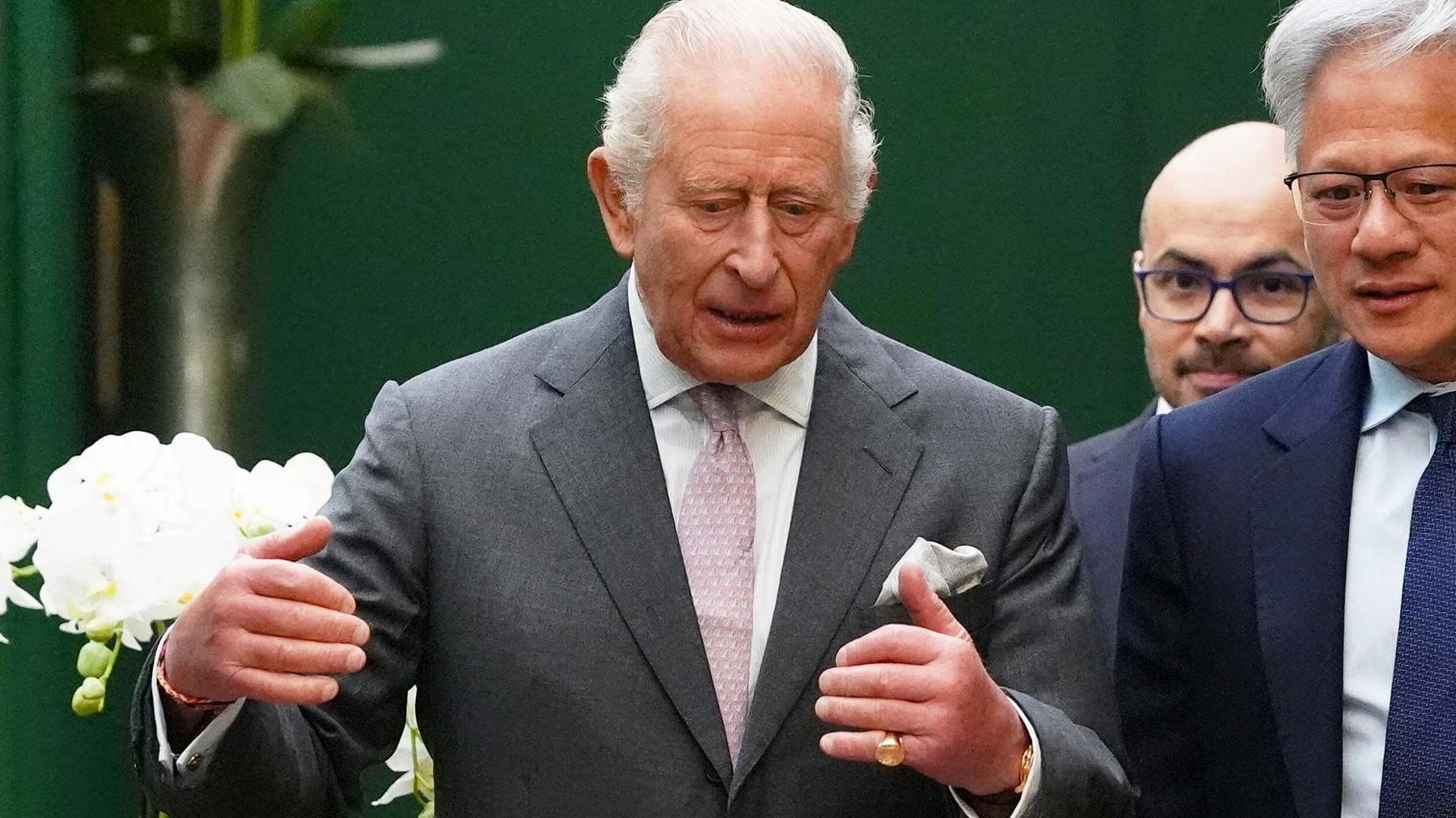 King Charles wearing a grey suit jacket and white shirt and pink tie, talking to Jensen Huang, the CEO of Nvidia, who is wearing a dark suit, white shirt and blue tie. Former Google DeepMind boss Sir Demis Hassabis is walking behind them
