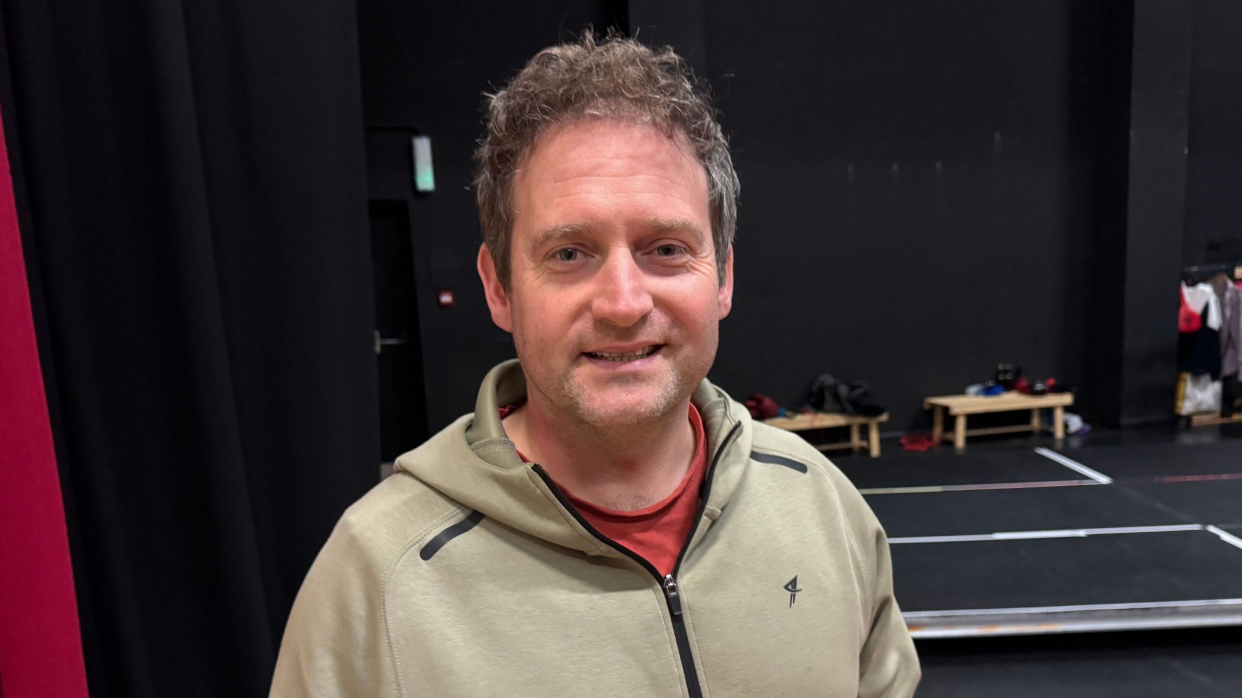 A man wearing a green jacket with a red T-shirt showing underneath. He is smilling.