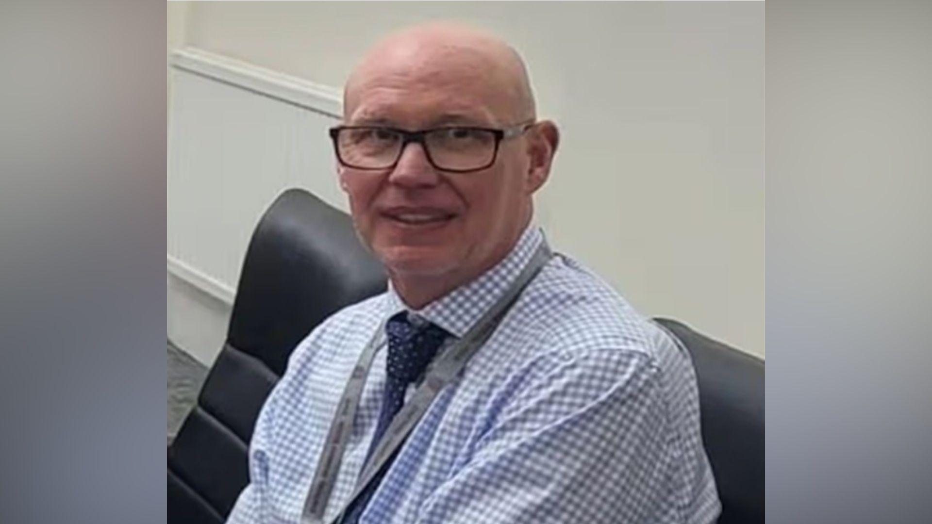 Michael Mullins, a bald man wearing dark-framed glasses, a blue-and-white checked shirt and a dark blue patterned tie, sits in a black office chair facing the camera.