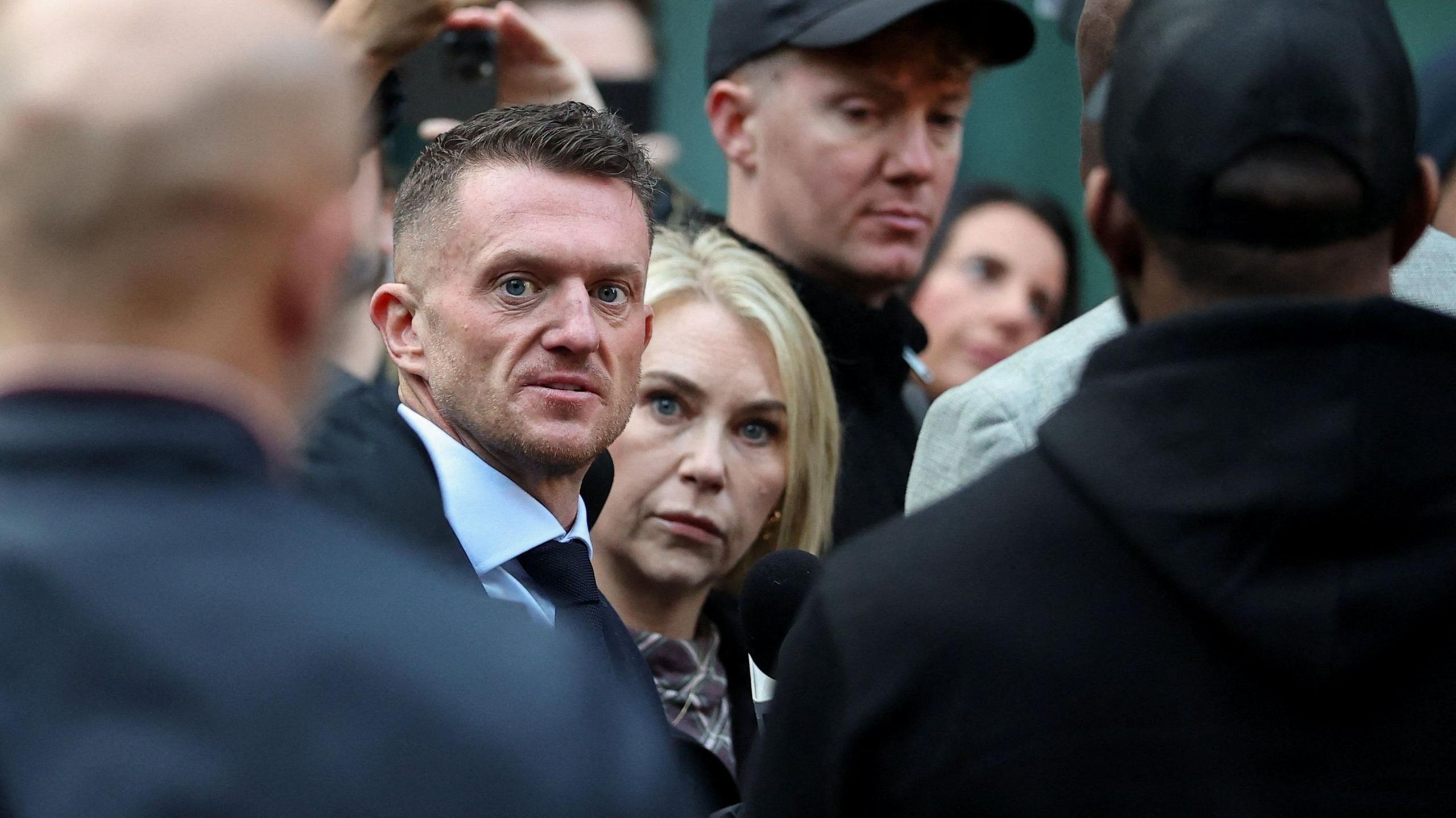 Tommy Robinson stares at the camera as he walks into court, surrounded by a close crowd and security officers. He is wearing a white shirt and black tie. Next to him is a woman with blonde hair and blue eyes, who also stares right at the camera.