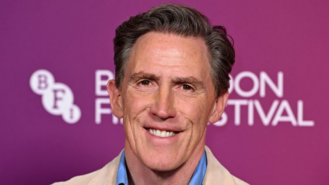 Rob Brydon at a screening of The Trip to the Northern Lights. Brydon wears a beige suit jacket and a light blue shirt. He is photographed from the shoulders up. He is smiling and looking at the camera.
