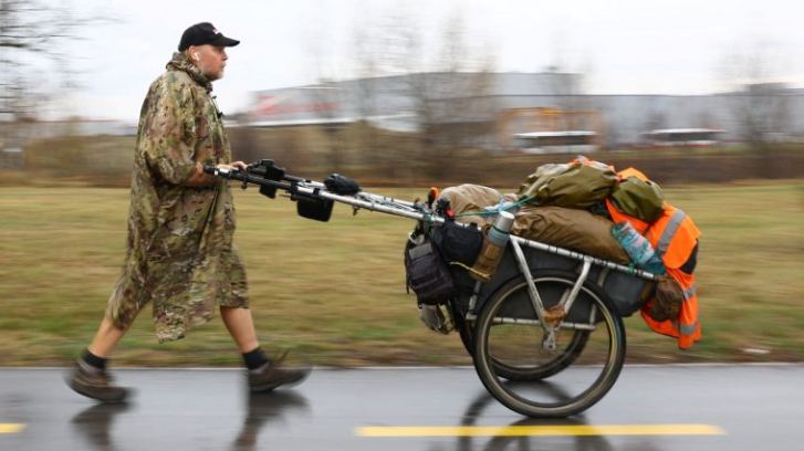 A man wearing a green camouflaged poncho, a baseball cap and boots walks along a wet road, pushing a two-wheeled trolley containing all of his possessions.