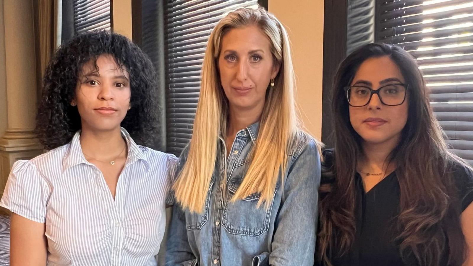 Three women holding neutral expressions as they pose for a photo. One of the women has black curly hair, another has straight blonde hair while the third, wearing glasses, has brown hair.