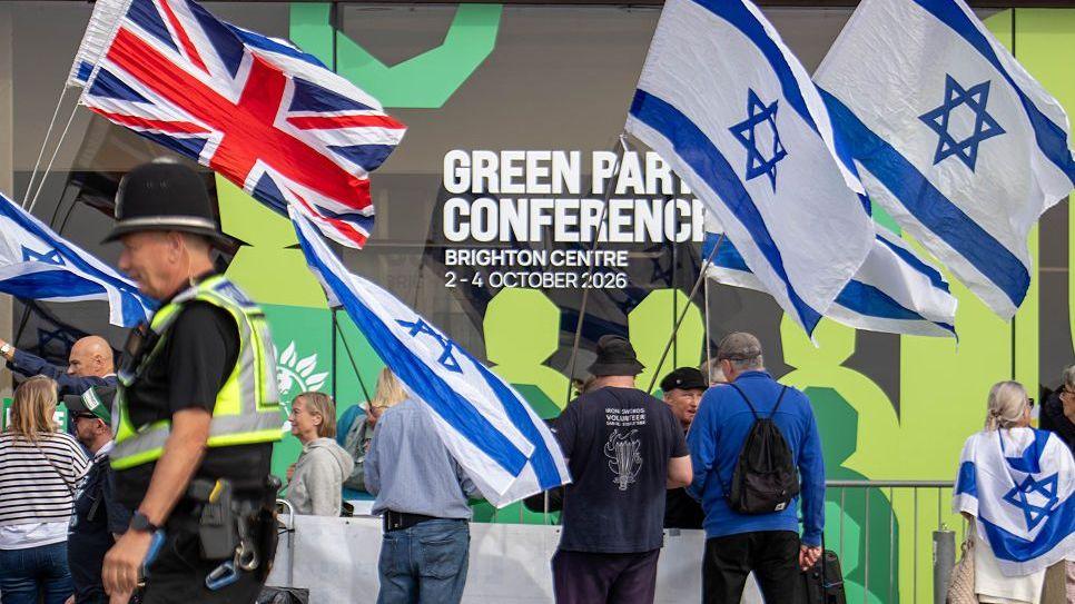 An image outside the Green Party conference of some people waving Israeli flags as well as a Union Jack. There is a policeman in the foreground.