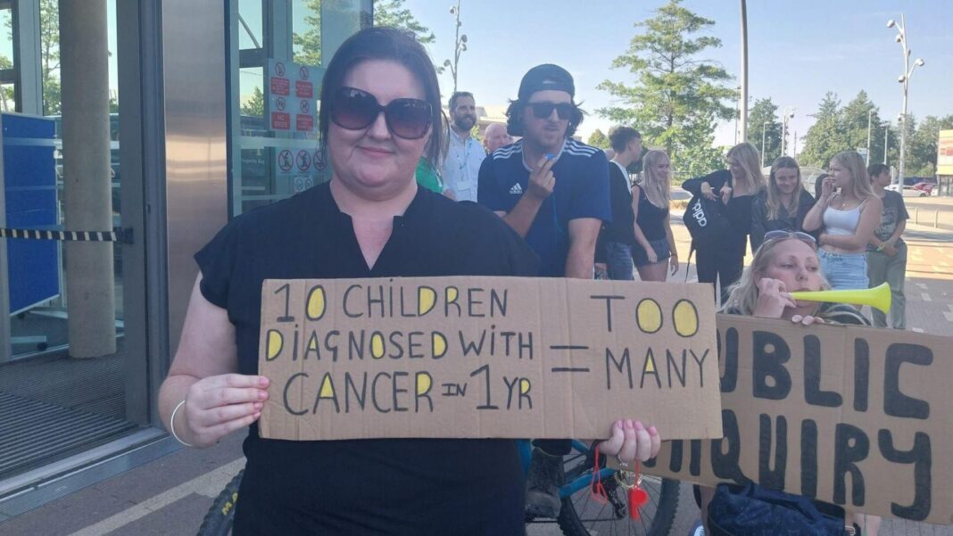 Council vows support for ‘Toxic Town’ public inquiry A woman is holding a cardboard banner saying too many children are being diagnosed with cancer. Another woman holds one calling for a public inquiry. Other people are gathered in the background.