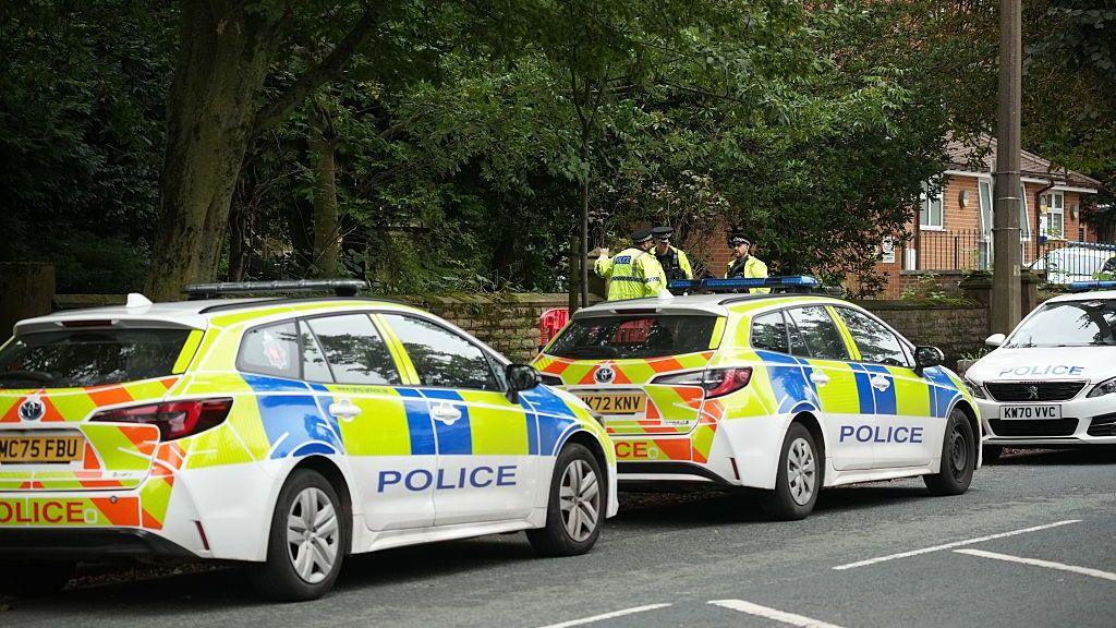 Two marked police cars parked at the side of a tree-lined road. Some officers wearing hi-vis yellow jackets are standing next to one of the cars