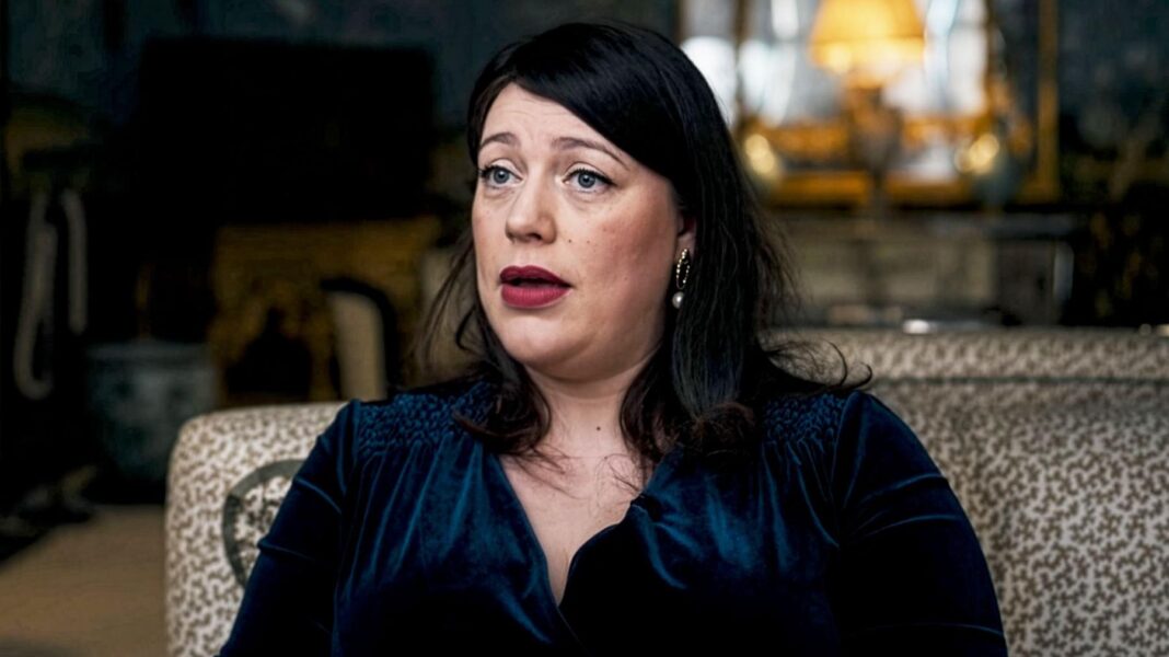 Head and shoulders shot of Alicia Kearns, who is wearing a midnightblue velvet top with pearl earrings and dark red lipstick. She sits on a pale, patterned sofa with a mirror and a lamp in the background.