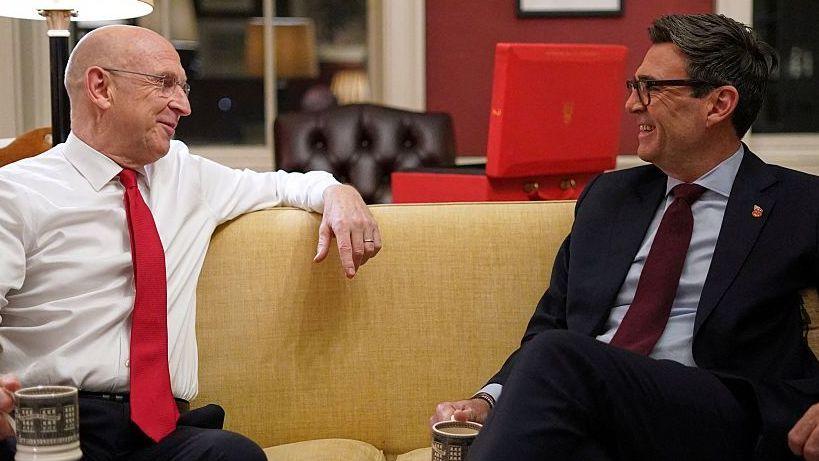 Chancellor of the Exchequer John Healey ahead of the UK budget at 11 Downing Street in London on October 26, 2026 sitting on a sofa smiling and talking to Prime Minister Andy Burnham. Both men are holding cups of tea.