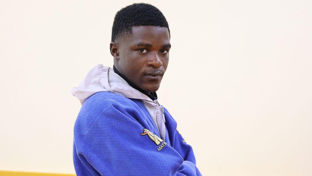 Judoka determined to prove refugees belong at Dakar 2026 Arno Yafunga Botay, visible from chest up wearing a light grey hoodie underneath a blue judo jacket, stands side on to the camera in front of a white wall and looks over his right shoulder into the lens with a determined expression