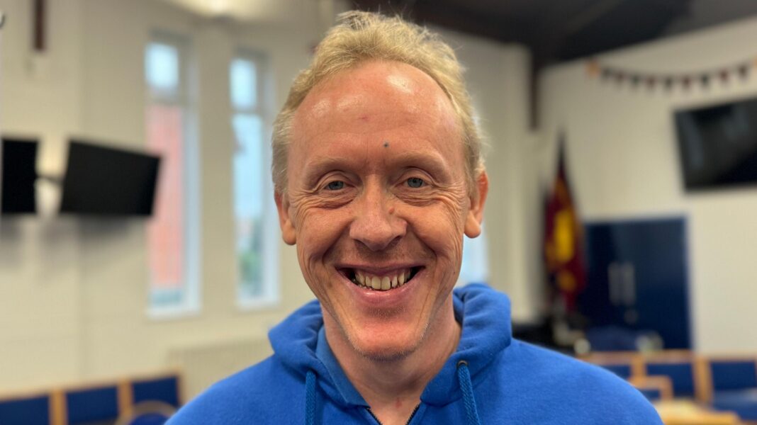 Music group with learning difficulties fulfils late MP’s Broadway dream David Stanley is smiling inside a classroom. He is wearing a blue hoody.