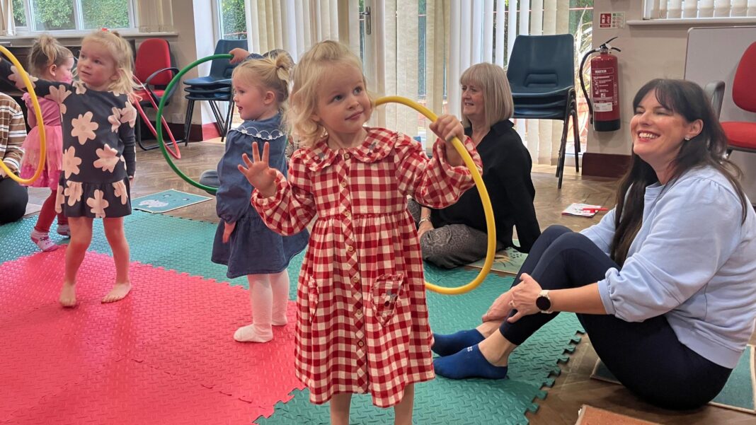 Potty training scheme to roll out to 20 more areas Four pre-school girls are dancing with different coloured hoops on a colourful mat in a church hall. They're being watched by their parents and grandparents who are sat on the mat next to them.