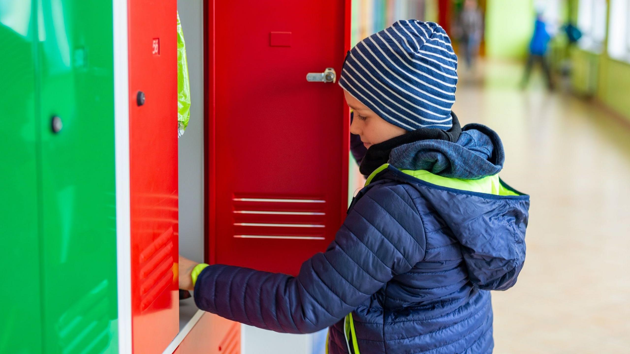 A child wearing a blue and white stripey hat and a blue anorak stands in front of a red school locker