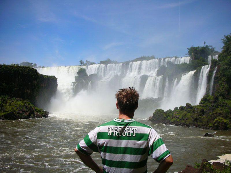 A young man with his back to the camera wearing the football strip while looking at a massive waterfall.