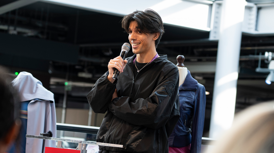 A picture of a man smiling holding a microphone. He wears a black top and has dark black hair.