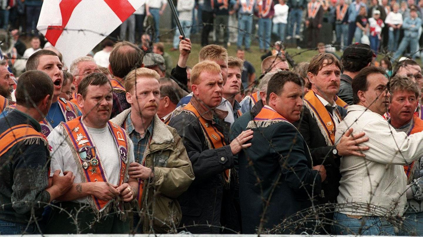 A crowd of men wearing orange sashes are standing n front of barbed wire. A St. George's Cross flag is being waved by a man.