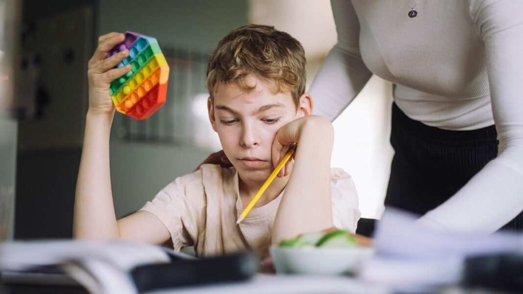 ADHD and autism at risk of over-diagnosis, says government review Young boy sits at table with pencil in one hand and colourful puzzle in the other. A woman stands over him.