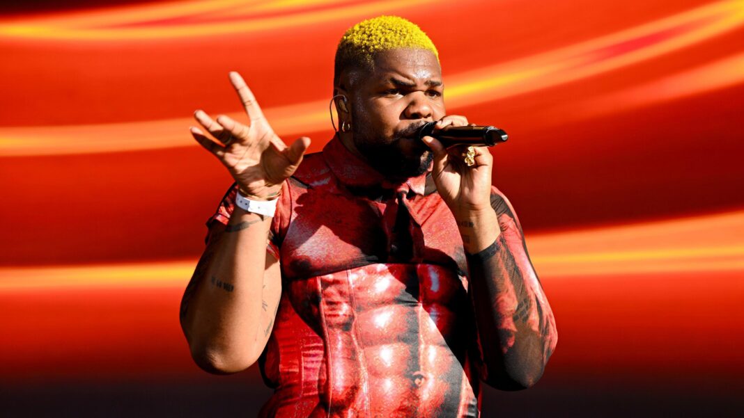 MNEK sings into a microphone while performing on stage. He wears a red top with a muscular body printed on it and has yellow hair.