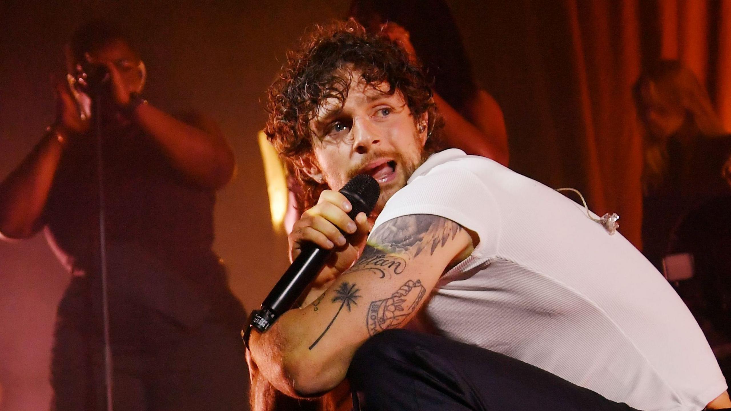 Tom Grennn is singing into a microphone while crouched on a stage with backing singers stood behind him