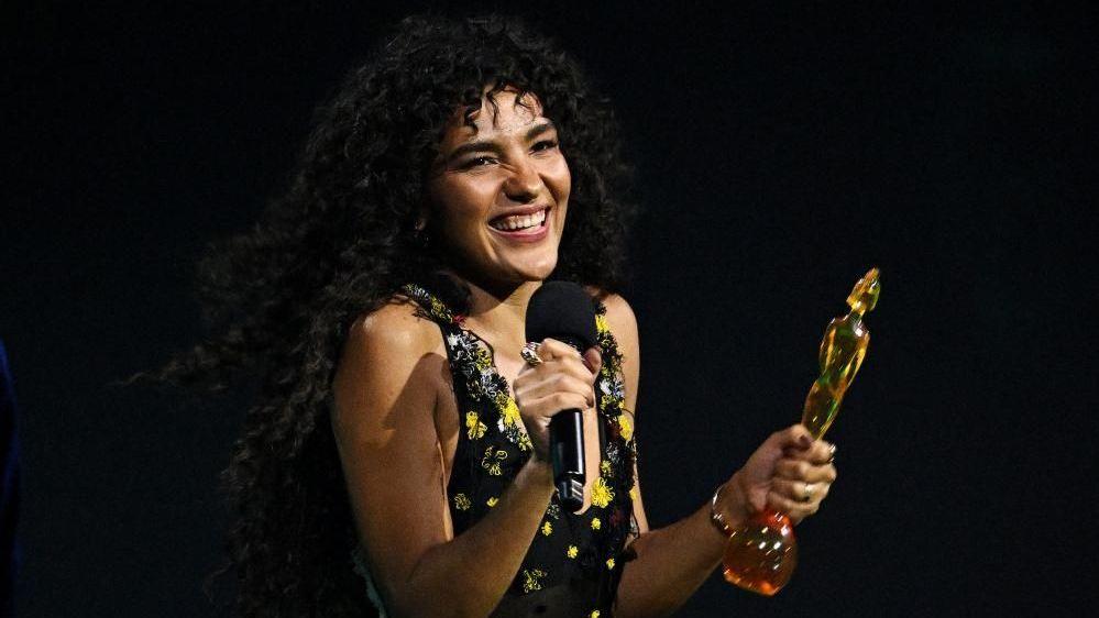 A woman with long curly black hair and wearing a black and yellow dress is smiling while holding a Brit award in her left hand and a microphone in her other hand.