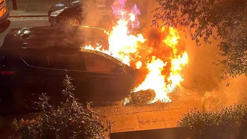 A black car engulfed in flames on a leafy north London street