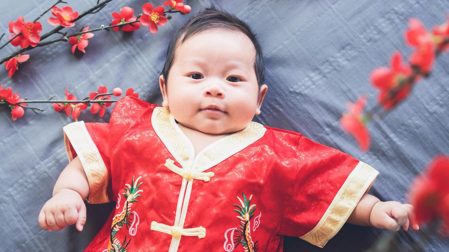 A baby lying down on a patterned grey cloth while dressed in a red traditional Chinese outfit with gold linings. Some red flowers surround him.