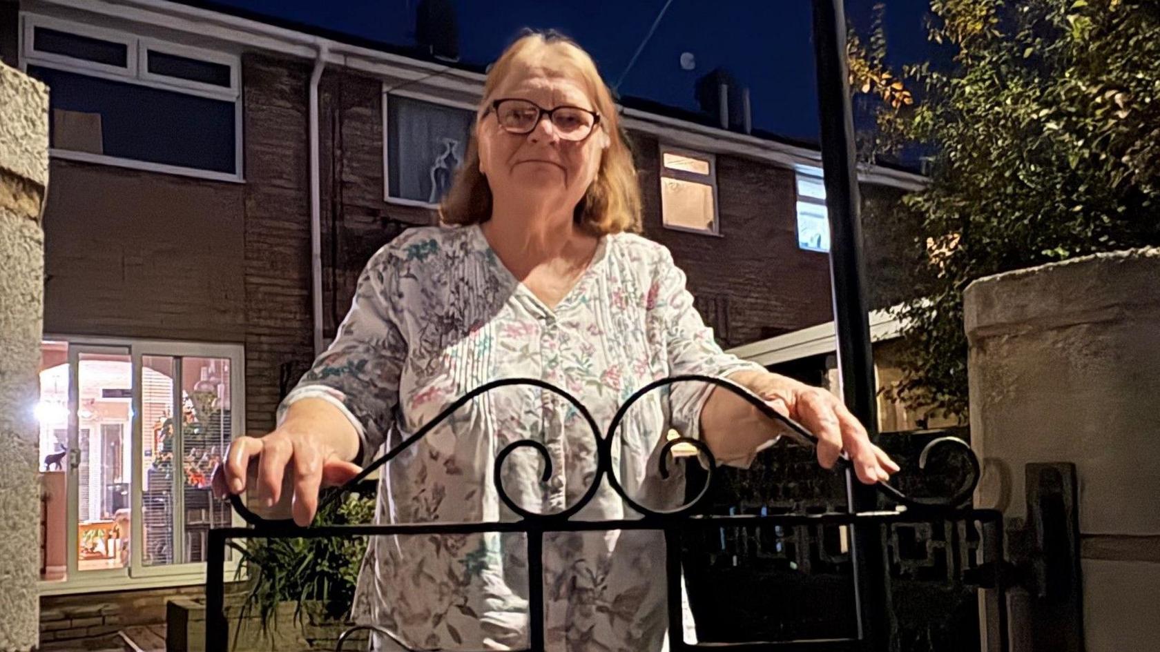 A bespectacled woman with greying light brown hair rests her hands on a wrought iron gate in front of her home. The photo is taken at night, with light coming from the living room behind her.